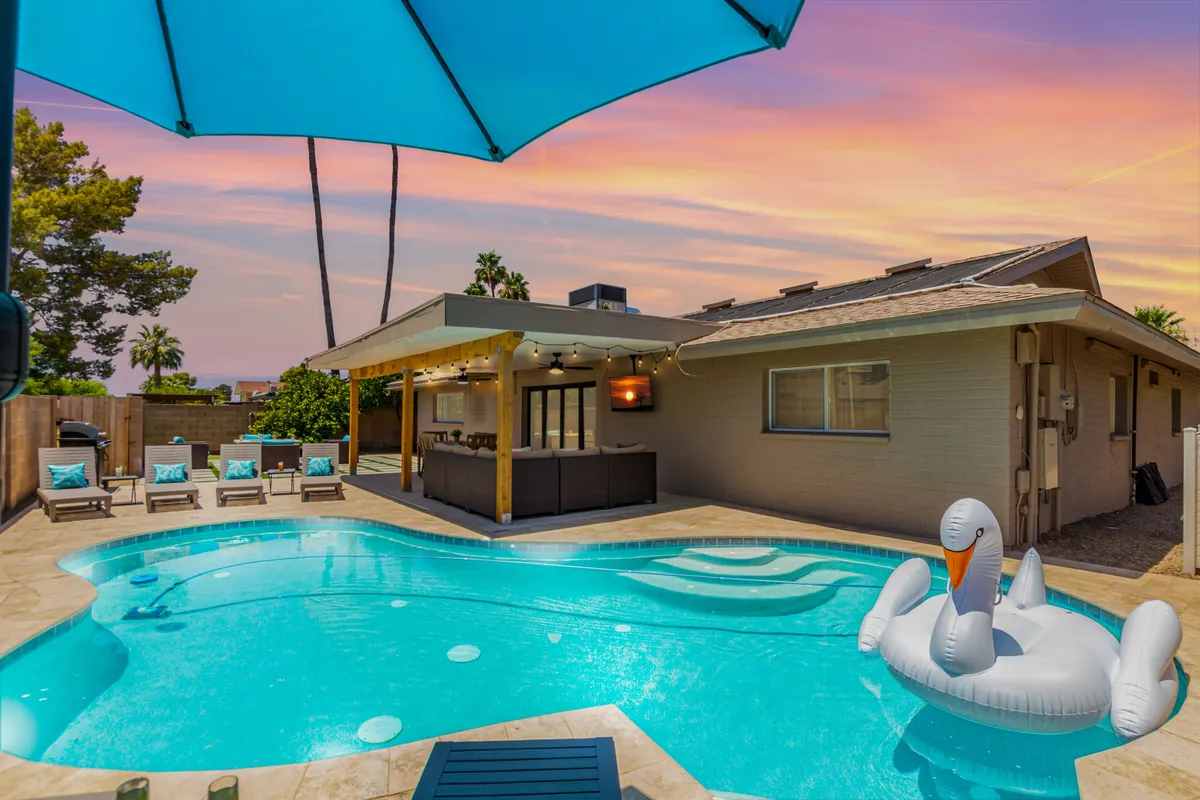 The hero image shows a beautiful backyard oasis featuring a sparkling swimming pool, a comfortable outdoor seating area shaded by a poolside umbrella, and a playful inflatable swan floating in the water. The scene is set for fun and relaxation, suggesting swimmable afternoons, lively social gatherings, and memorable sunset moments.