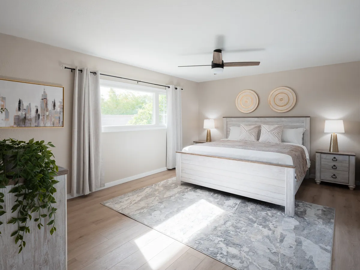 A serene bedroom with a comfortable bed set against a window that lets in soft natural light. Artwork, a houseplant, and minimal decor complete the soothing, inviting environment ideal for reading or a peaceful night's sleep.
