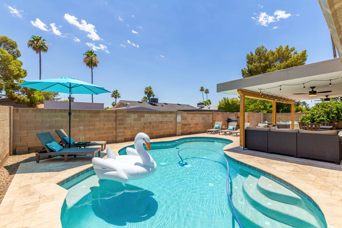 A vibrant exterior setting featuring a sparkling swimming pool with a playful swan float, an inviting outdoor seating area under a wide umbrella, and the lush presence of palm trees, all set against a sunny Scottsdale sky.