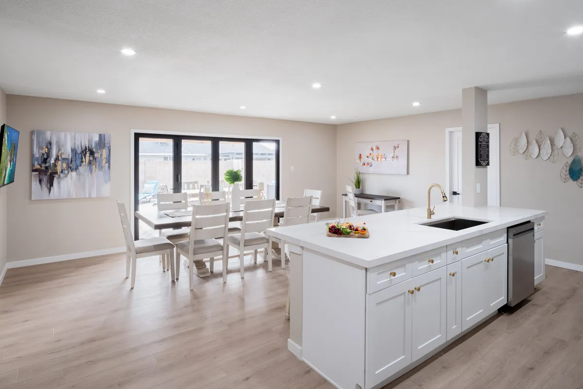 An inviting open-concept space featuring a kitchen island, a dining table, decorative wall art, and large sunlit windows. The layout connects the kitchen and dining areas, with natural light pouring in, showcasing the harmonious blend of function and style ideal for shared meals and gatherings.