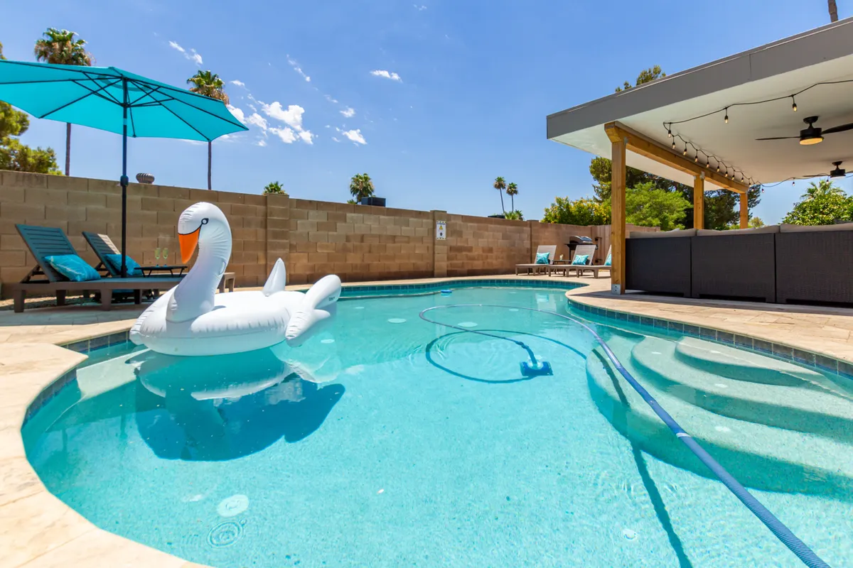 A sun-drenched backyard scene features a sparkling swimming pool at the center, surrounded by an inflatable swan float, lounge chairs, a large umbrella, and an inviting outdoor seating area. The area is set for playful afternoons and relaxing moments, with hints of sunset color in the sky.
