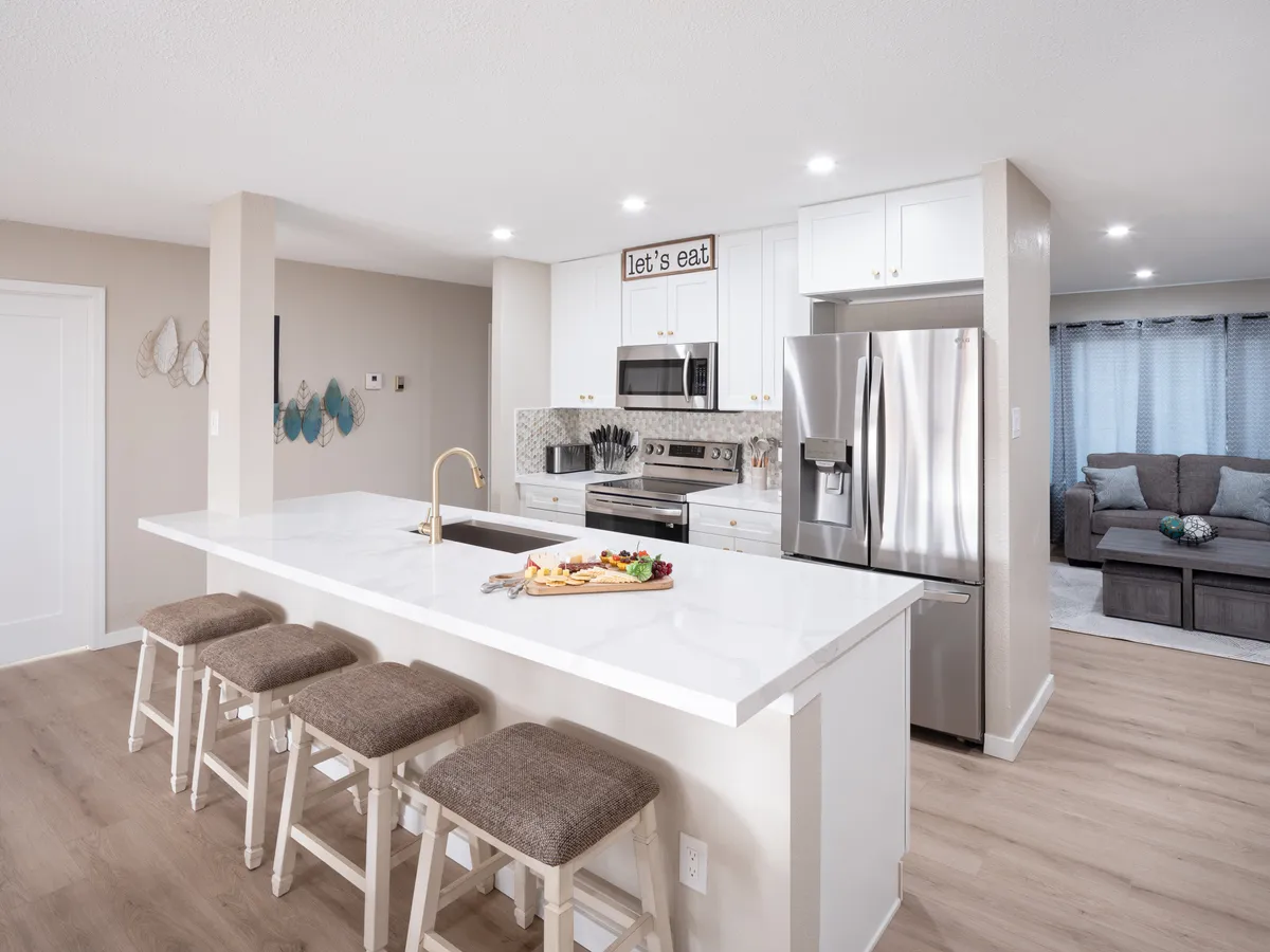 A warm, welcoming kitchen scene with a central island surrounded by stools, modern appliances visible in the background (including stainless steel finishes), a decorative sign, and a food platter on the counter. The space feels ready for both cooking and socializing, with thoughtful touches that evoke family gatherings.