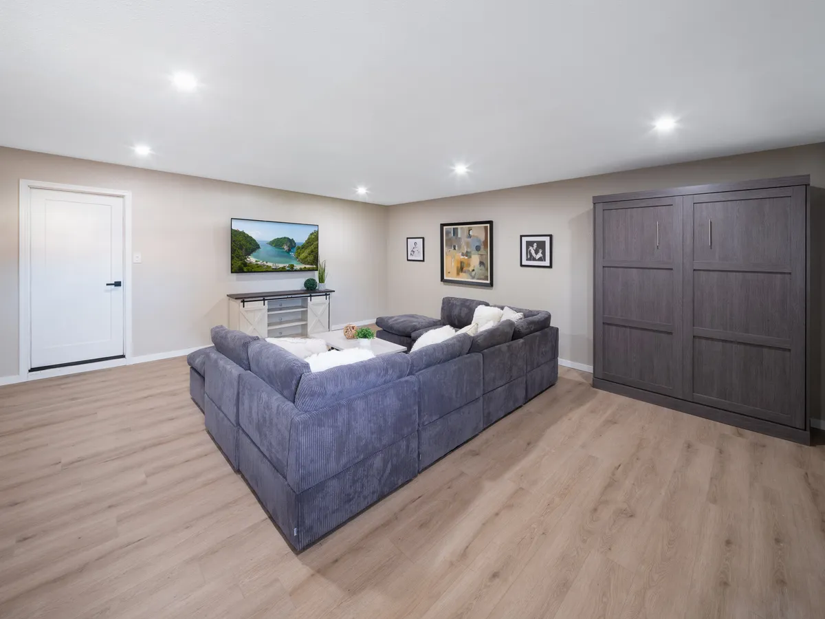A spacious living room featuring a large grey sectional sofa, a substantial TV displaying a serene nature scene, colorful artwork on the walls, and a storage cabinet. The setup is ideal for group activities such as movie and game nights, with ample comfortable seating encouraging social interaction.