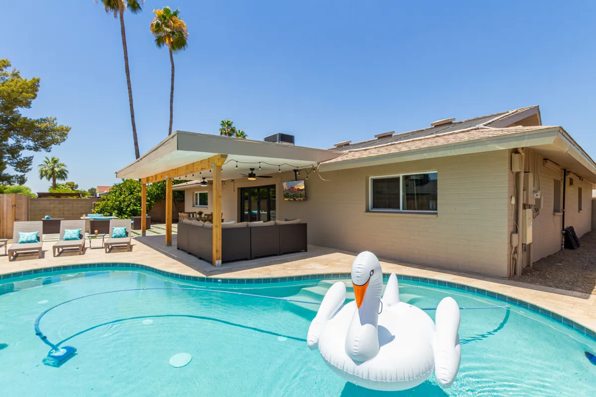 A sunlit backyard view with a sparkling swimming pool, a playful inflatable swan, and an inviting outdoor seating area surrounded by palm trees. The scene radiates a fun, leisurely atmosphere ideal for families enjoying time together after an adventurous day.