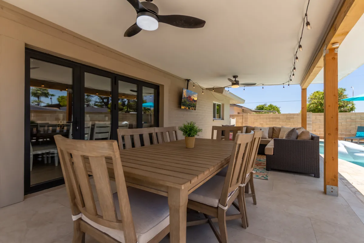 A welcoming outdoor dining and lounge area with a table, comfortable seating, a pool in the background, and a ceiling fan overhead. The space is set up for both relaxation and socializing, with views to the tranquil yard and pool.